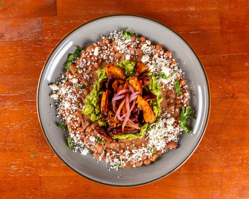 A gray plate of refried beans and whole beans topped with guacamole, roasted plantains, and pickled red onions.