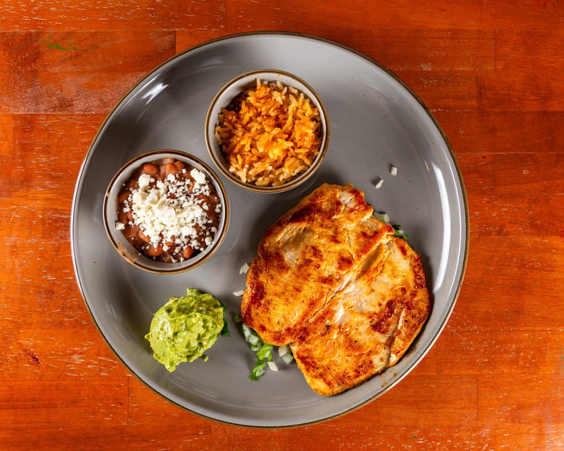 Grilled chicken breast with sides of rice, beans topped with cheese, and guacamole on a gray plate.