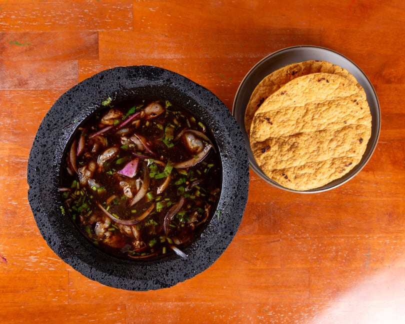 Molcajete bowl of dark soup with shrimp, red onions, and cilantro, served with tortillas.