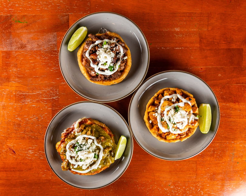 Three sopes on gray plates with various toppings, sour cream, cheese, and lime wedges on a wooden table.