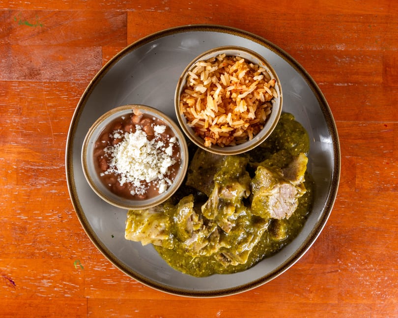 Pork in green chile sauce with rice and beans topped with cheese on a gray plate.