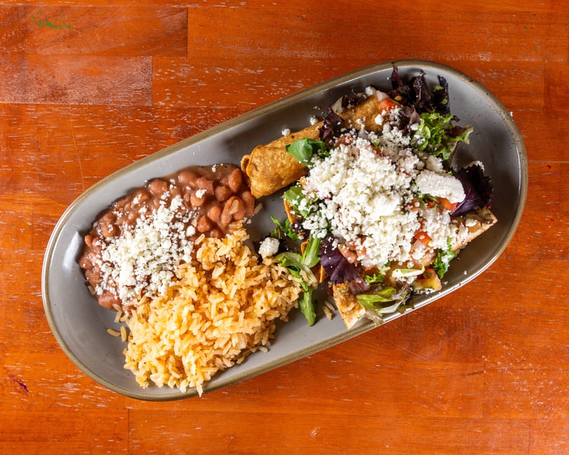 A plate of Mexican food with rice, beans, a taquito, and a tostada salad, all topped with cheese.