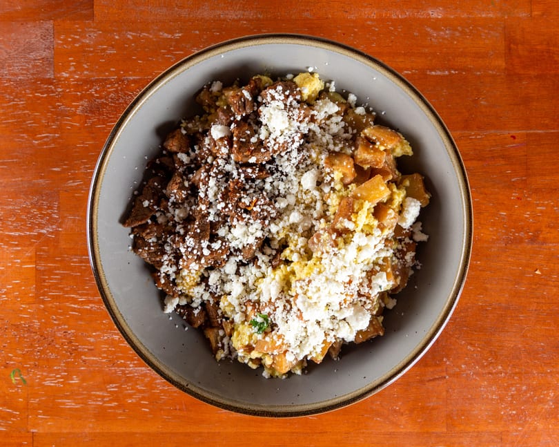 Overhead view of a bowl with scrambled eggs, diced potatoes, steak, and crumbled cheese.