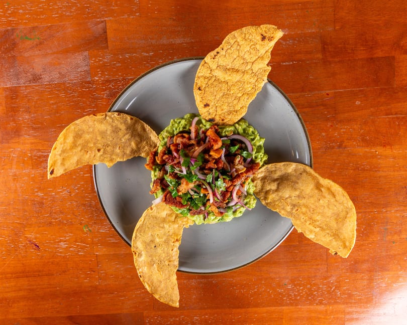 Guacamole topped with chicharron, red onion, and cilantro, surrounded by tortilla chips.