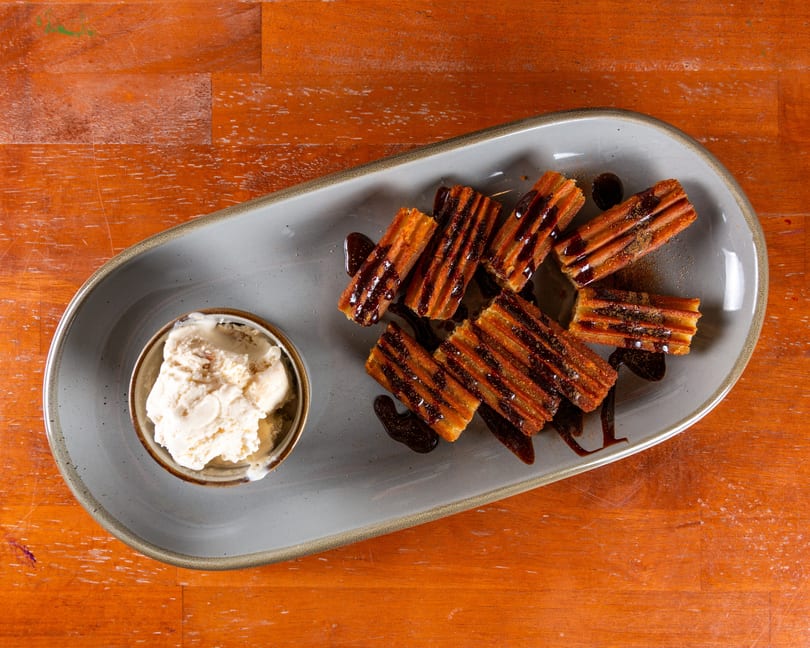 Churros drizzled with chocolate sauce and a side of vanilla ice cream on a gray plate.