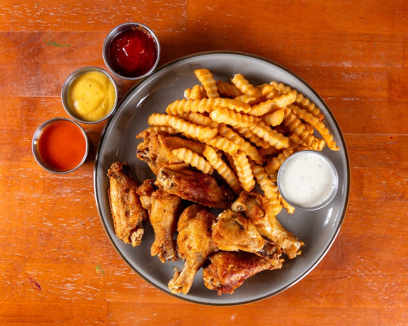 Crispy fried chicken wings and crinkle-cut fries with ketchup, mustard, hot sauce, and ranch on a plate.