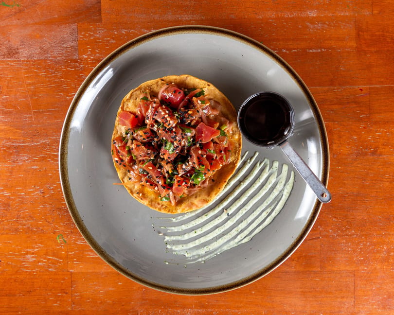 Tuna tostada with sesame seeds, cilantro, and red onion, served with a side of sauce.