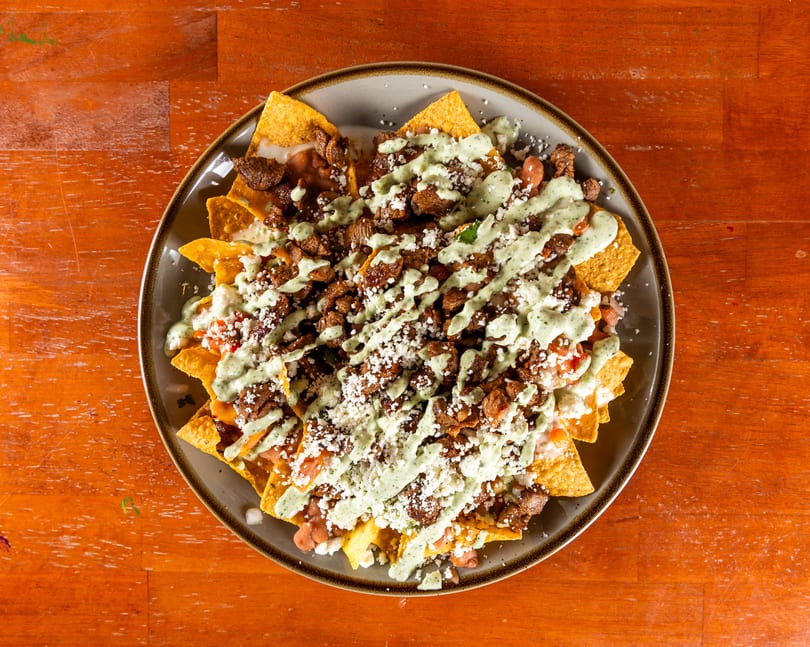Overhead view of a plate of nachos with meat, beans, cheese, and green sauce on a wooden table.